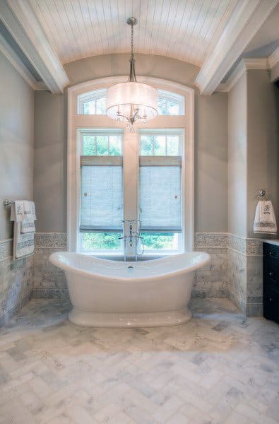 Elegant bathroom with a freestanding bathtub, arched window, and hanging chandelier; marble floor and detailed walls