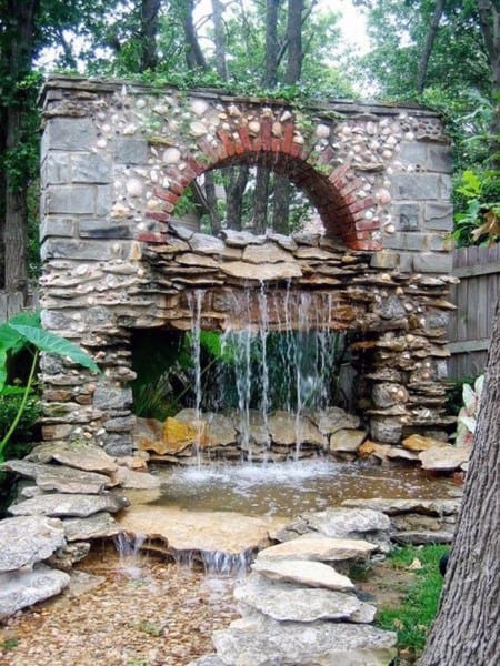 Stone arch fountain with cascading water flowing into a small pond surrounded by rocks.