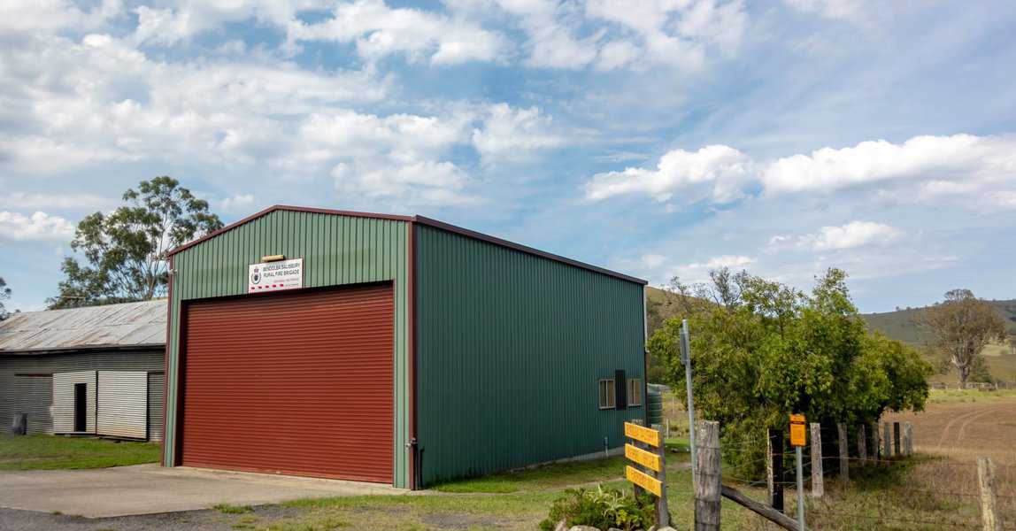 Large green aluminum shed with a red roller door in a rural setting surrounded by fields.