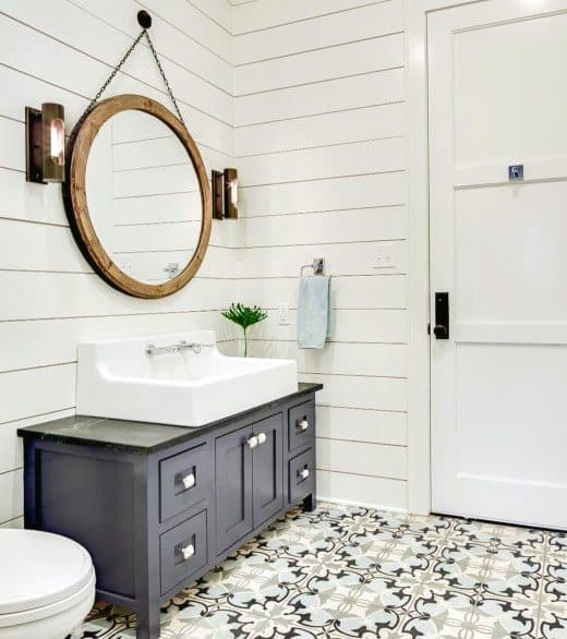 Half bath with shiplap walls, a round mirror, black vanity with white sink, and geometric tile flooring