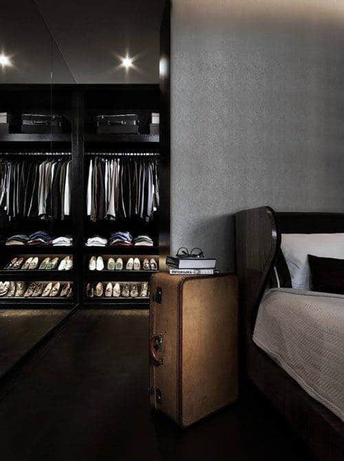 Modern bedroom with open closet showing clothes and shoes, suitcase-style bedside table, and dark decor