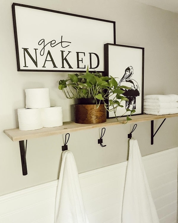 Wooden bathroom shelf with towel hooks, potted plant, toilet paper rolls, and framed wall art.