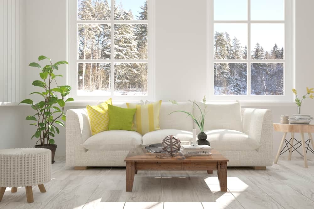 Bright Scandinavian living room with white furniture, green accents, a wooden coffee table, and large snowy window views