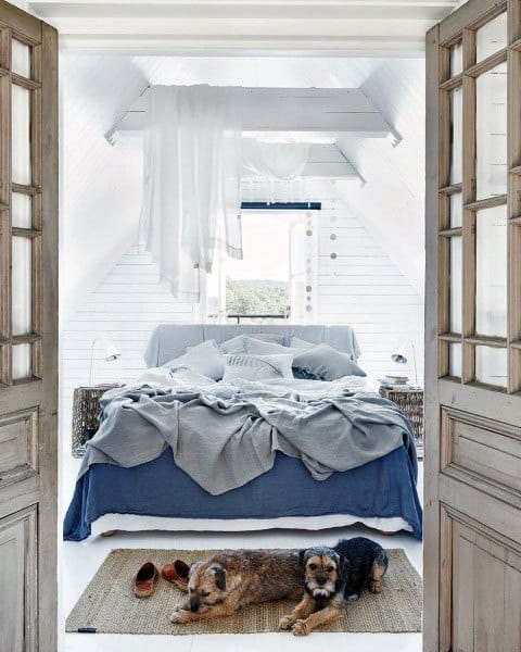 Cozy attic bedroom with an unmade bed, light blue bedding, and two small dogs lying on a rug near wooden doors