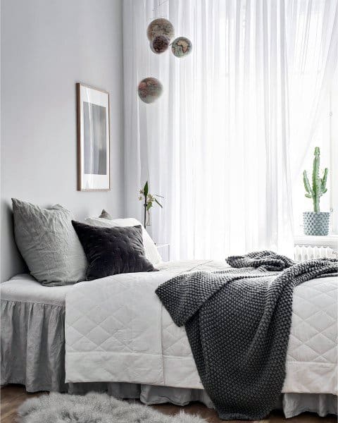 Minimalist bedroom with a neatly made bed, grey pillows, a knit throw, and a cactus by a sheer-curtained window