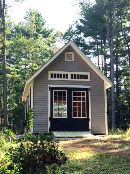 rey cottage-style shed with double doors in a wooded backyard environment.