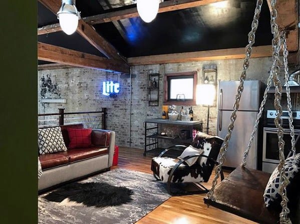 Industrial-style mancave with exposed beams, leather sofa, swing seating, and rustic brick walls.