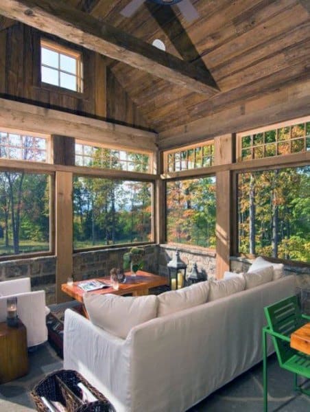 Cozy rustic sunroom with white couches, wooden ceiling, and scenic forest views.