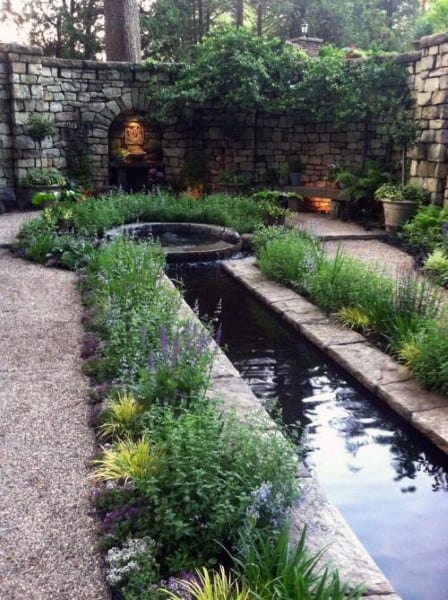 Long narrow stone pond with lush greenery and a stone wall backdrop in a peaceful garden space.