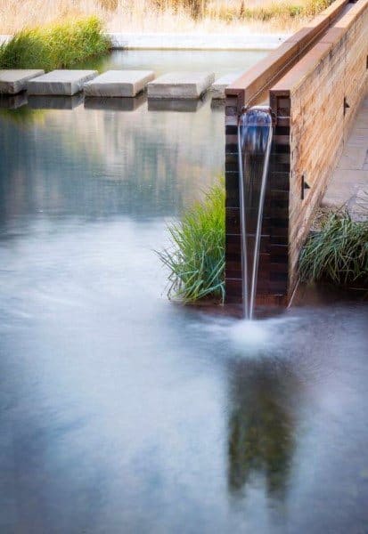 Modern waterfall feature with water flowing from a wooden wall into a serene pond, surrounded by plants.