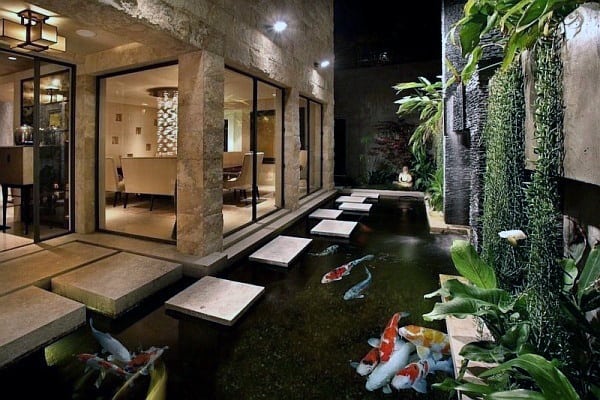 Koi pond with floating stone steps, lush greenery, and modern glass windows reflecting soft lighting.