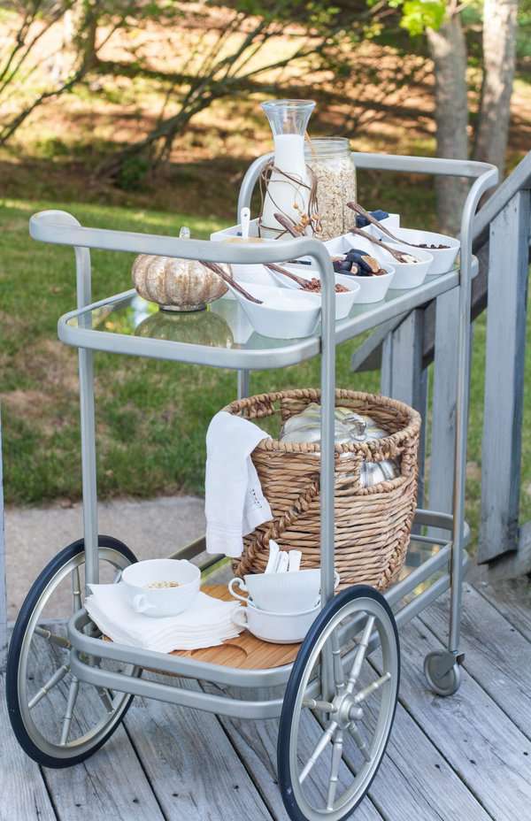 Gray bar cart with large wheels, serving bowls, a milk pitcher, and a wicker basket for outdoor dining.