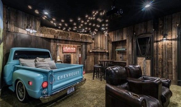 Rustic farmhouse mancave with vintage truck bed sofa, leather chairs, wood walls, and string lights.