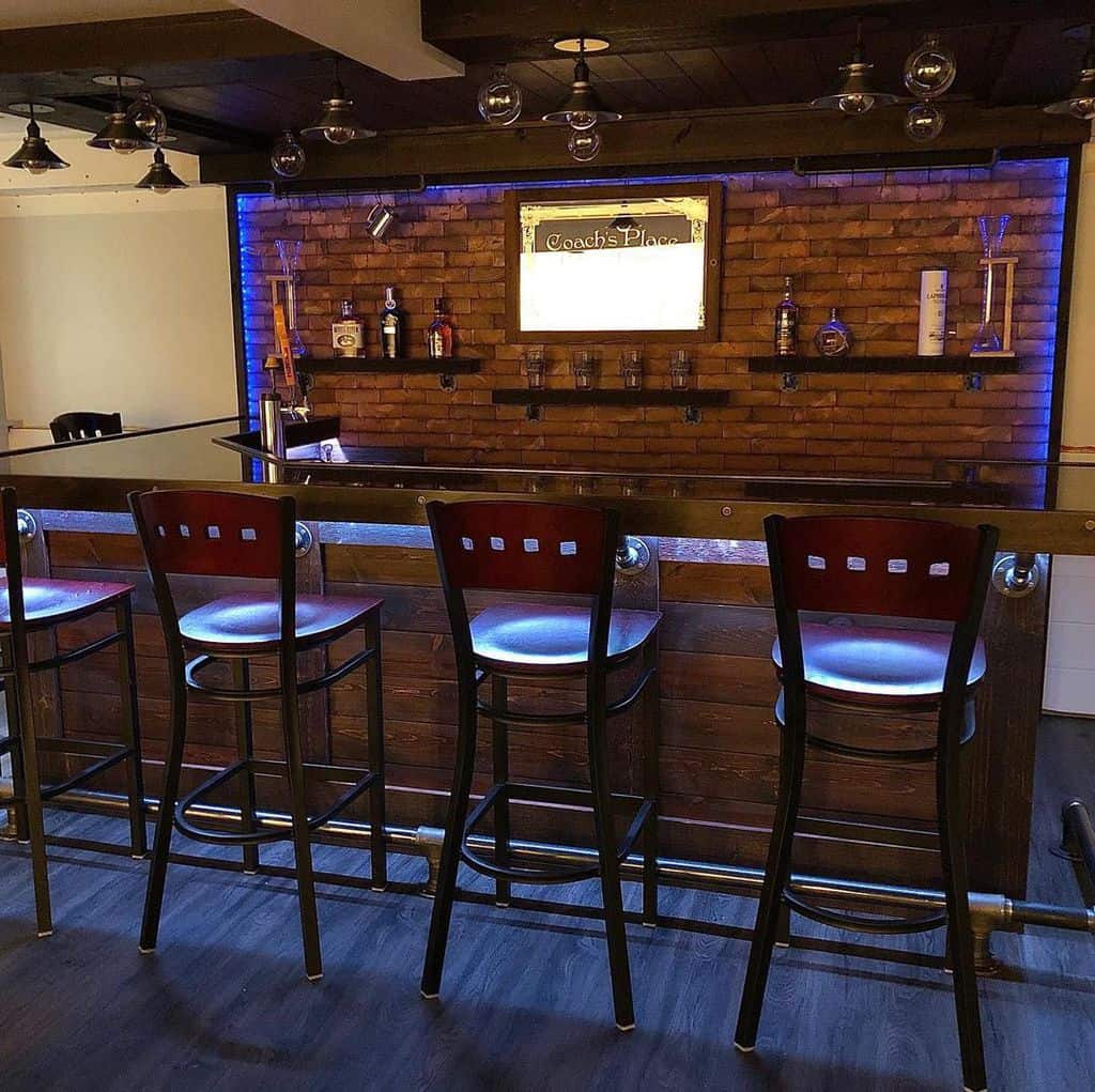 Brick basement bar with glowing blue lighting, wooden stools, and decorative shelves.