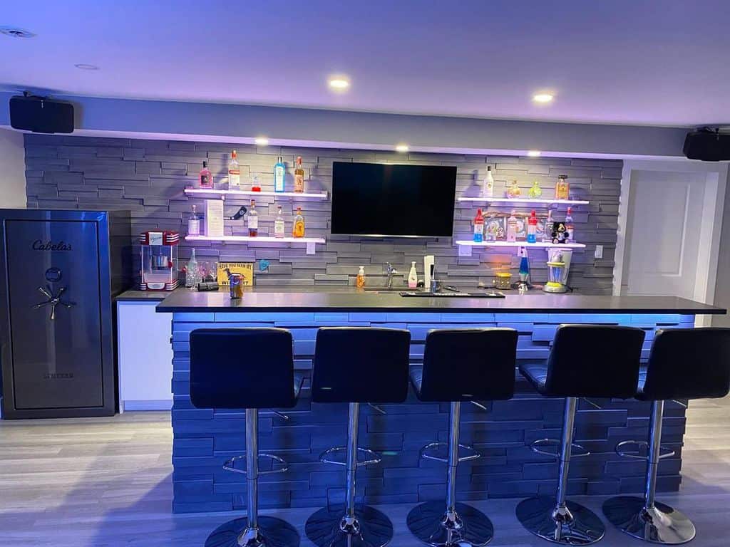 Contemporary basement bar with stone wall, illuminated shelves, bar stools, and flat-screen TV.