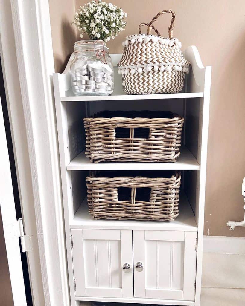 White shelving unit with wicker baskets and glass jar of flowers for bathroom storage.