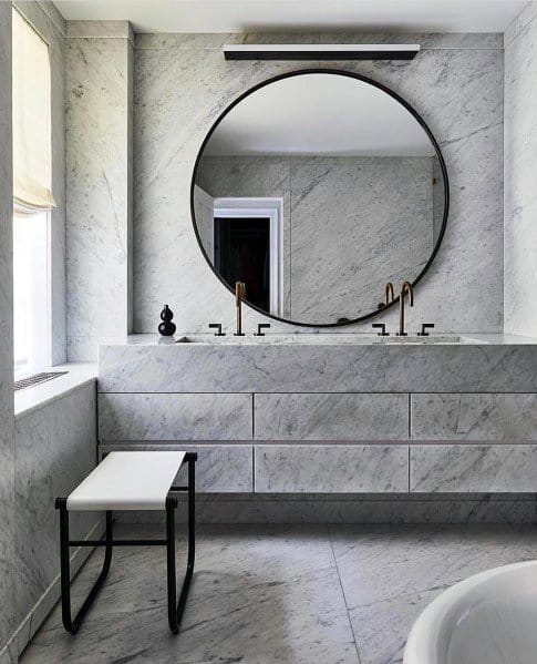 Modern bathroom featuring large round mirror, marble vanity, and a white bench.