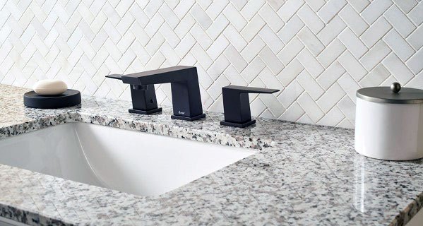 Bathroom with white herringbone tile backsplash, granite countertop, and black faucet.