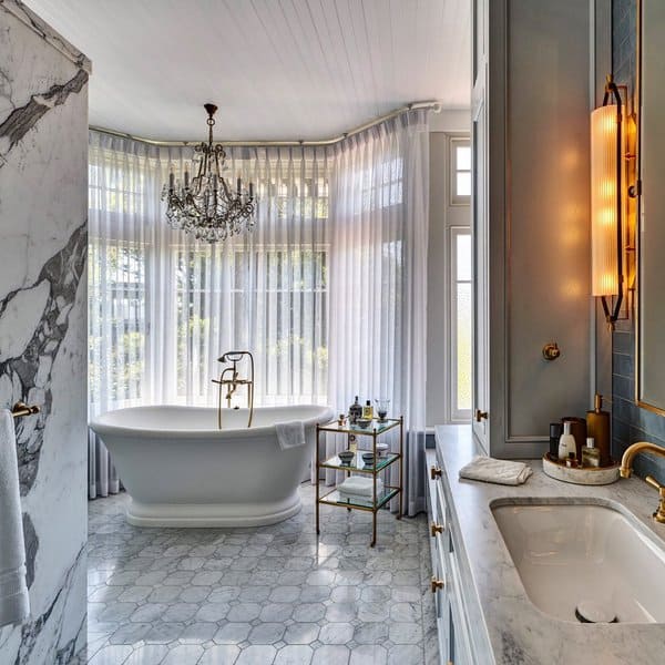 Freestanding bathtub with chandelier, marble accents, and sheer curtains in an elegant bathroom.