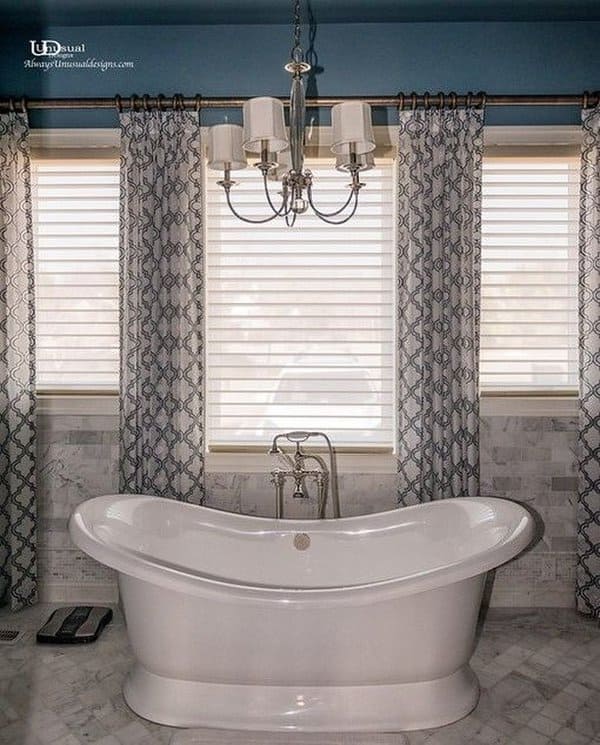 Freestanding bathtub with chandelier, patterned curtains, and marble tile flooring.