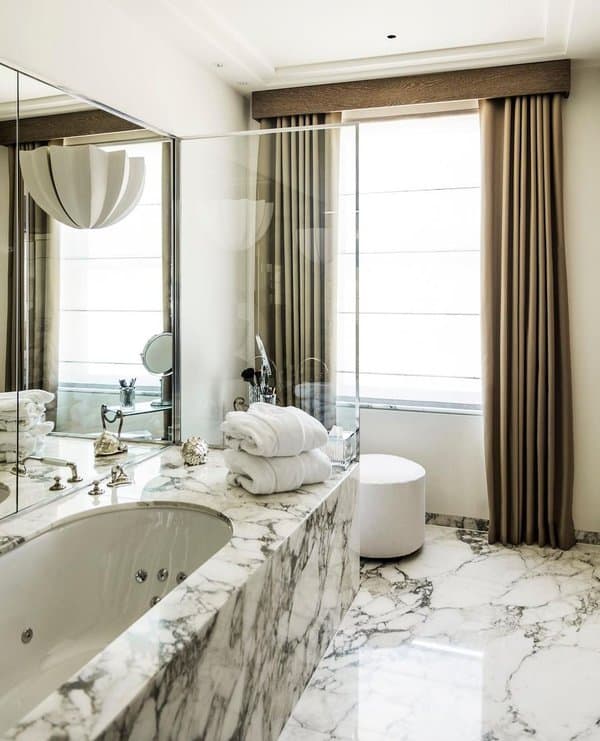 Luxurious bathroom with marble surfaces, large mirrors, and floor-to-ceiling beige curtains.