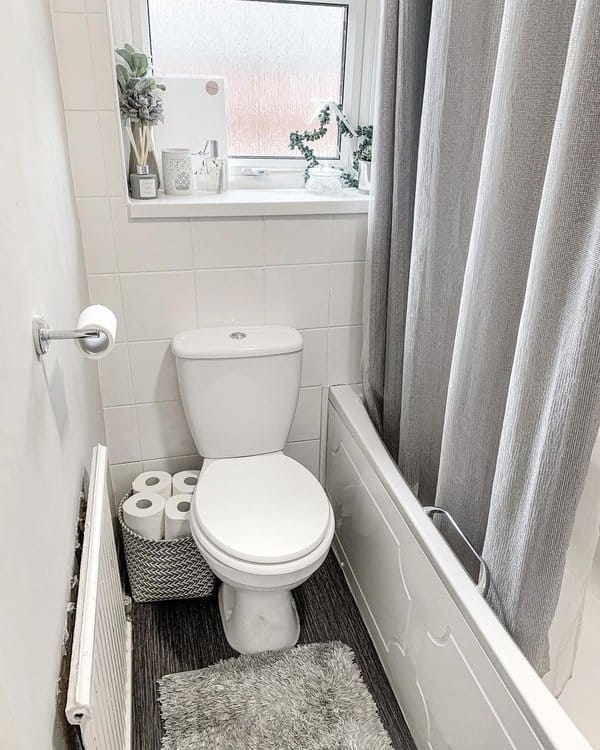 Small bathroom with toilet, gray curtain, basket of toilet paper, and window shelf decor..