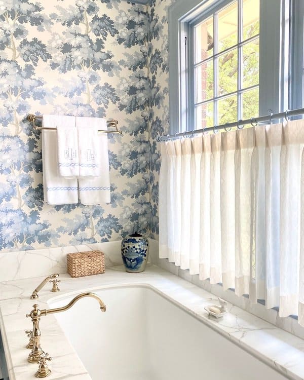 Blue floral wallpaper bathroom with a marble tub, white curtains, and brass fixtures.