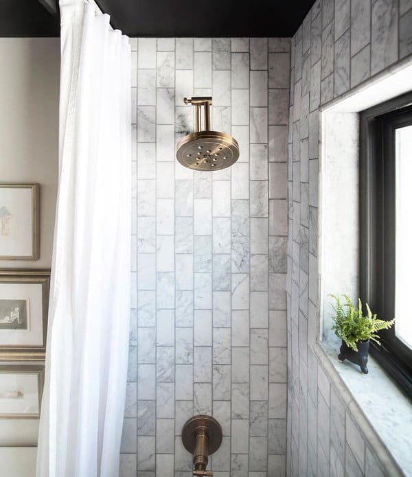 Marble-tiled shower with brass fixtures, white curtain, and small window with a plant.