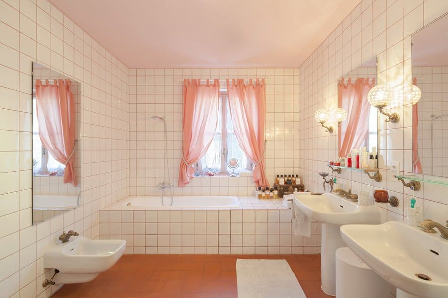 Vintage bathroom with pink curtains, white tiled walls, and dual sinks with brass fixtures.