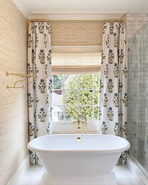 Freestanding tub with floral curtains, large window, and brass fixtures in a bright bathroom.