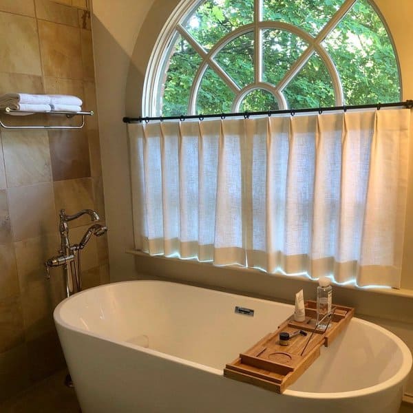 Freestanding tub with half-curtained arched window and wooden bath tray in a cozy bathroom.