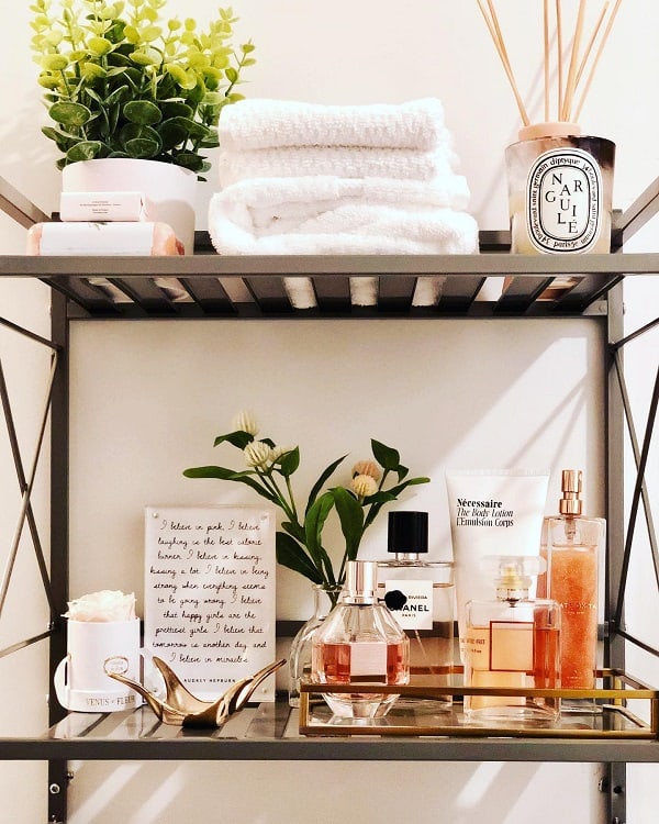 Metal bathroom shelf with folded towels, potted plant, perfumes, and decorative tray.