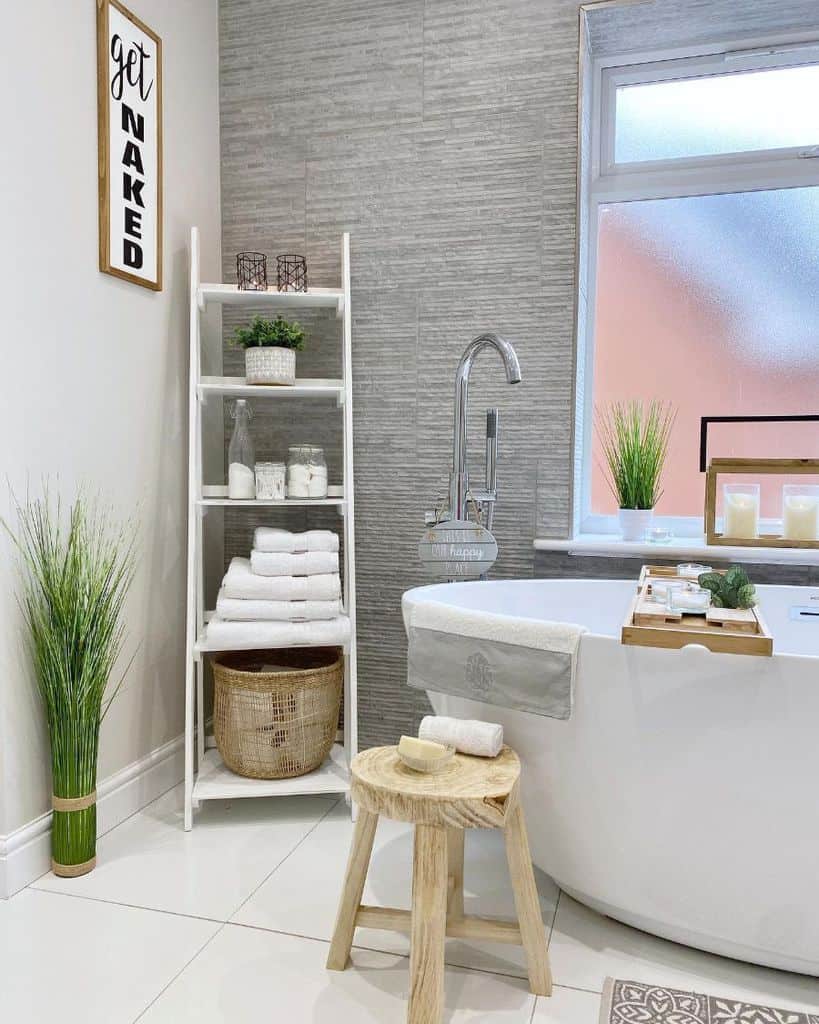 White bathroom storage rack with folded towels, wicker basket, and decor next to freestanding bathtub.