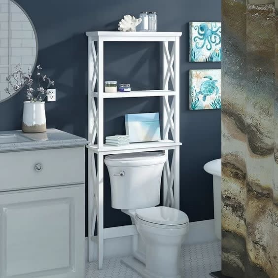 White over-the-toilet storage rack with shelves holding decor and towels in a navy blue bathroom.