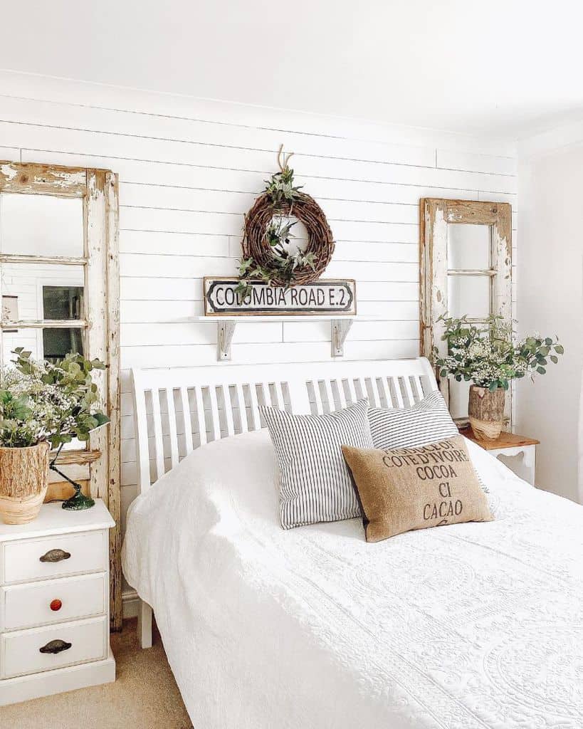 Bright bedroom with a white bed, rustic mirrors, pillows, and plants, adorned with a sign and wreath above the white headboard