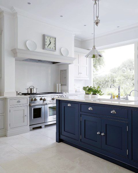 Bright kitchen with navy island, white cabinets, beige tile flooring, and stainless steel appliances, featuring natural light