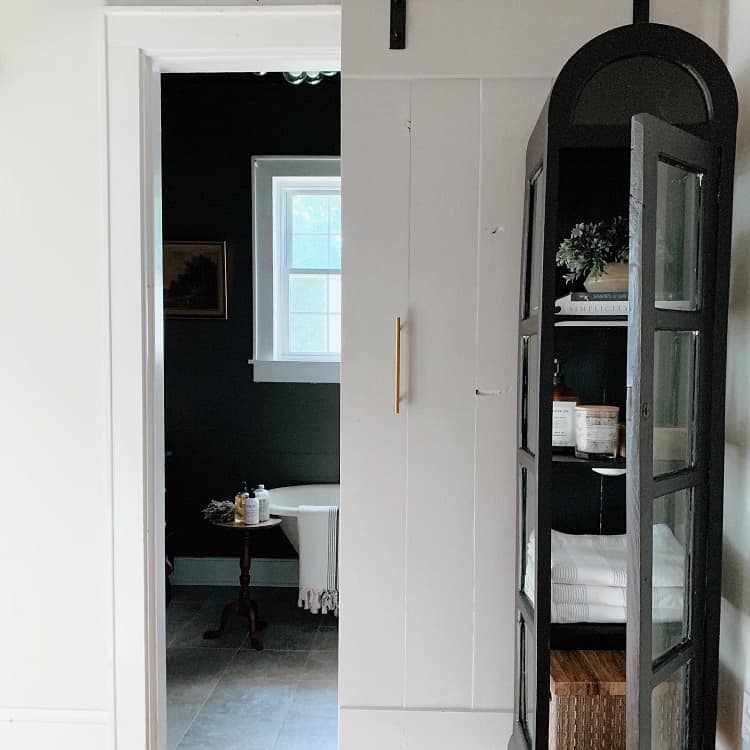 Black arched bathroom shelving unit with glass doors, next to a sliding barn door.