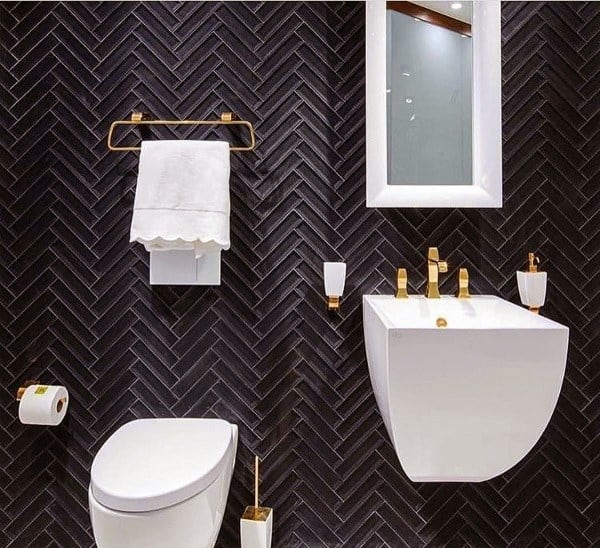 Modern bathroom with black herringbone tiles, white fixtures, gold accents, and a wall-mounted sink and toilet