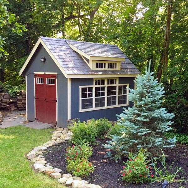 Blue backyard shed with red farmhouse-style doors and a shingled roof in a landscaped garden.