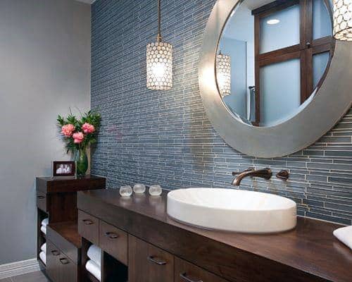 Wood vanity with round mirror, blue tile backsplash, and pendant lighting.