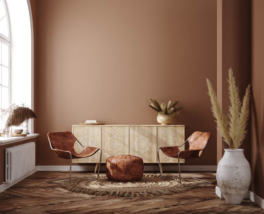 Cozy room with two brown leather chairs, a pouf, wooden sideboard, dried plants, and natural light from a large window