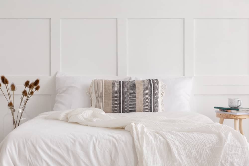 Minimalist white bedroom with a bed, striped cushion, dried plants in a vase, and a wooden side table with books and a mug