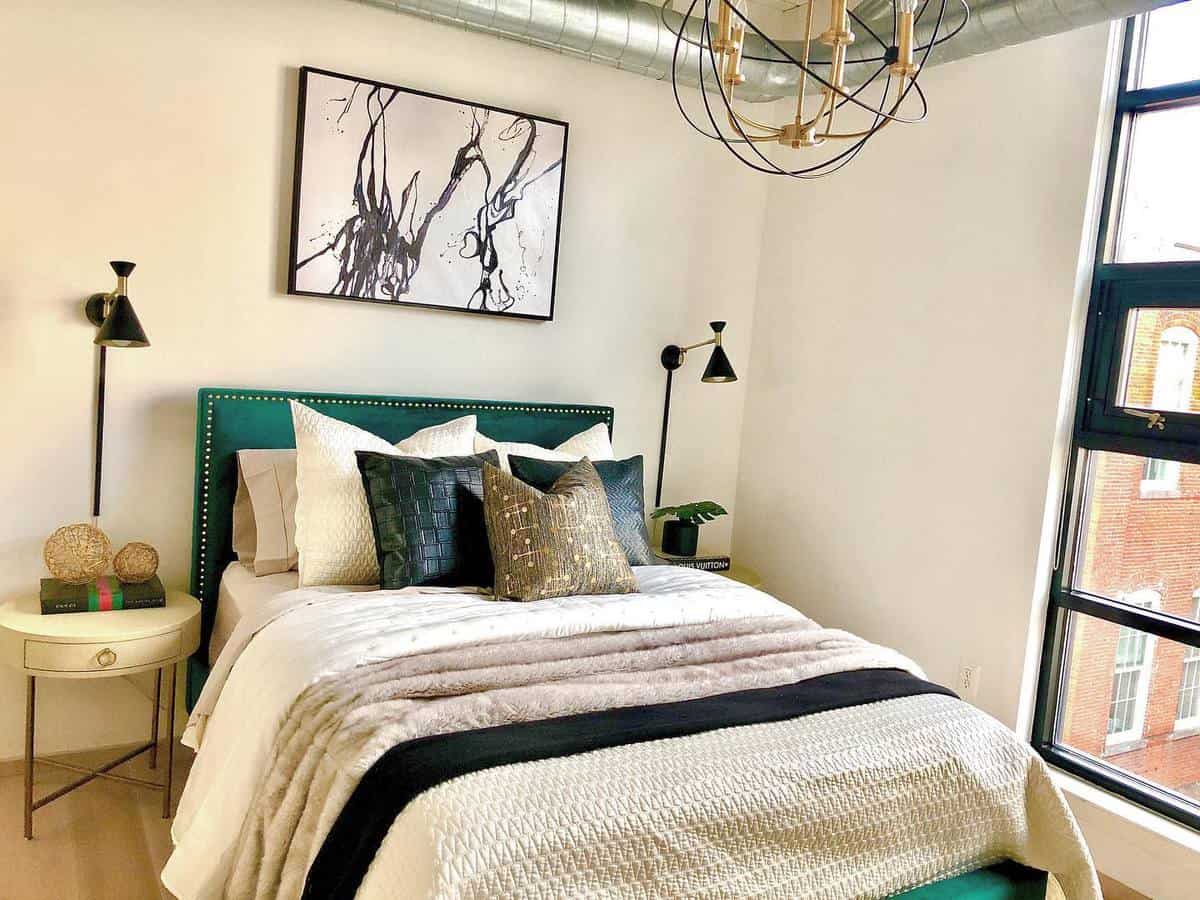 Modern bedroom with a green bed, abstract artwork, and large window. Decor includes pillows and a round side table