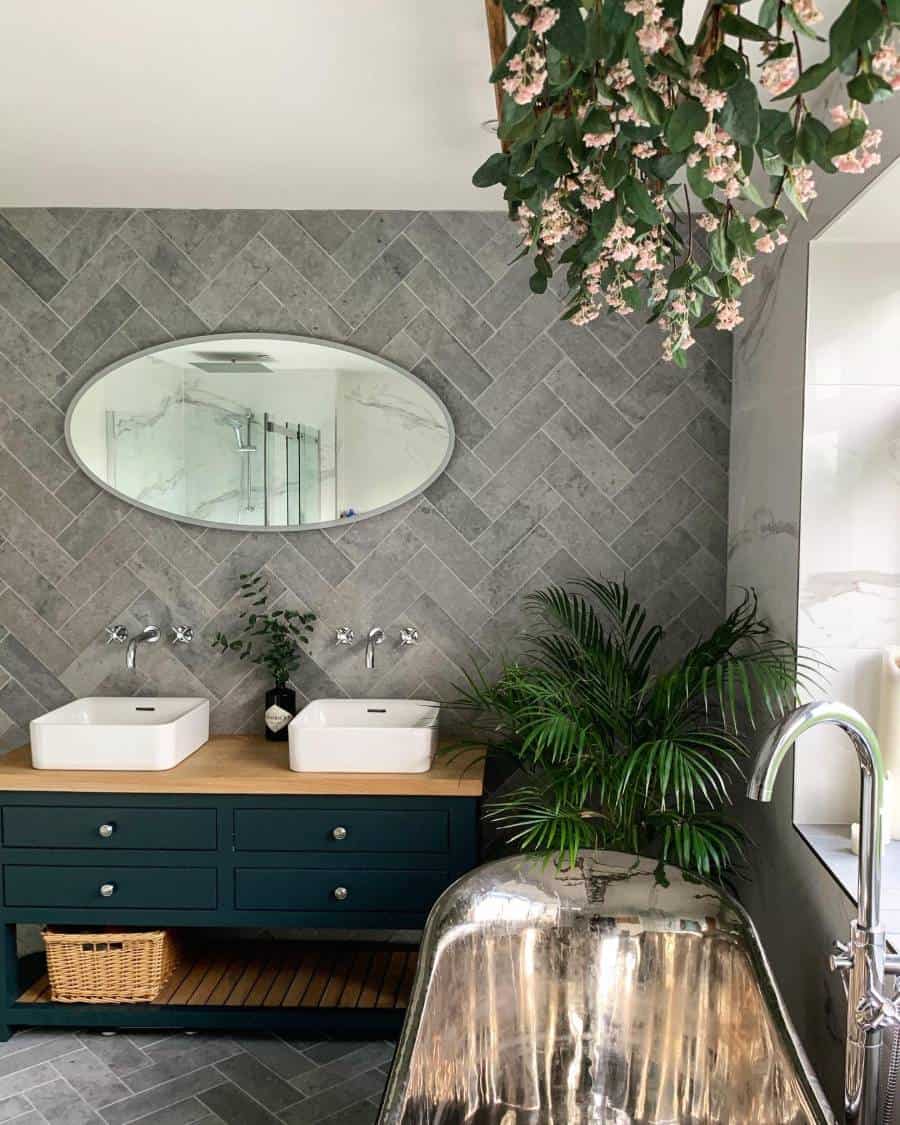 Modern bathroom with silver bathtub, double sinks, herringbone wall tiles, and hanging plants.