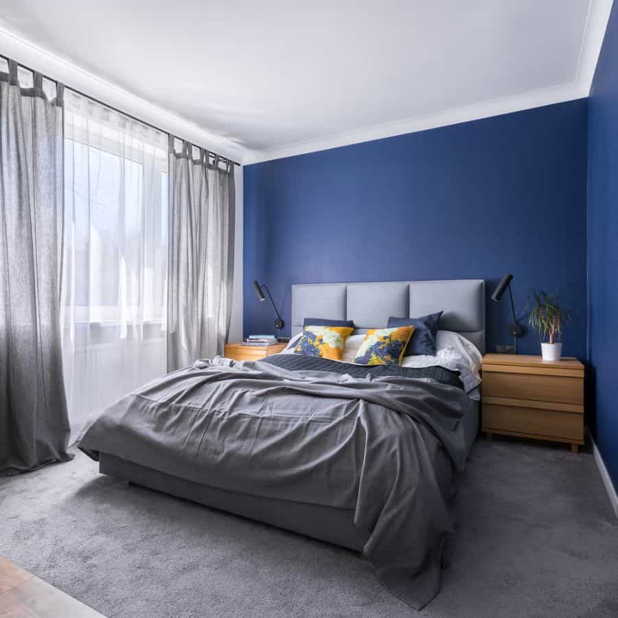 Modern blue bedroom with grey carpet, light headboard, and wooden nightstands.