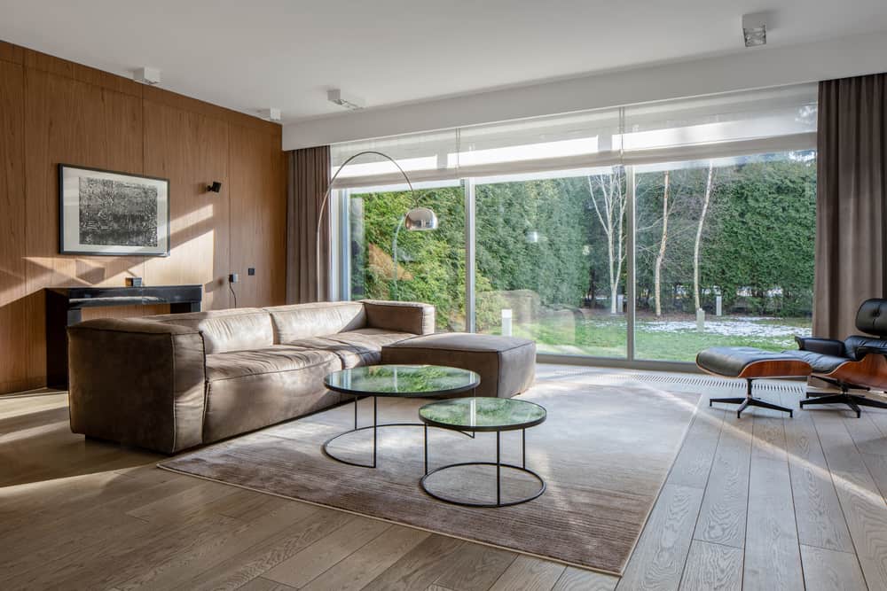 Contemporary living room with a brown leather sectional, glass coffee tables, wood-paneled walls, and large garden-facing windows