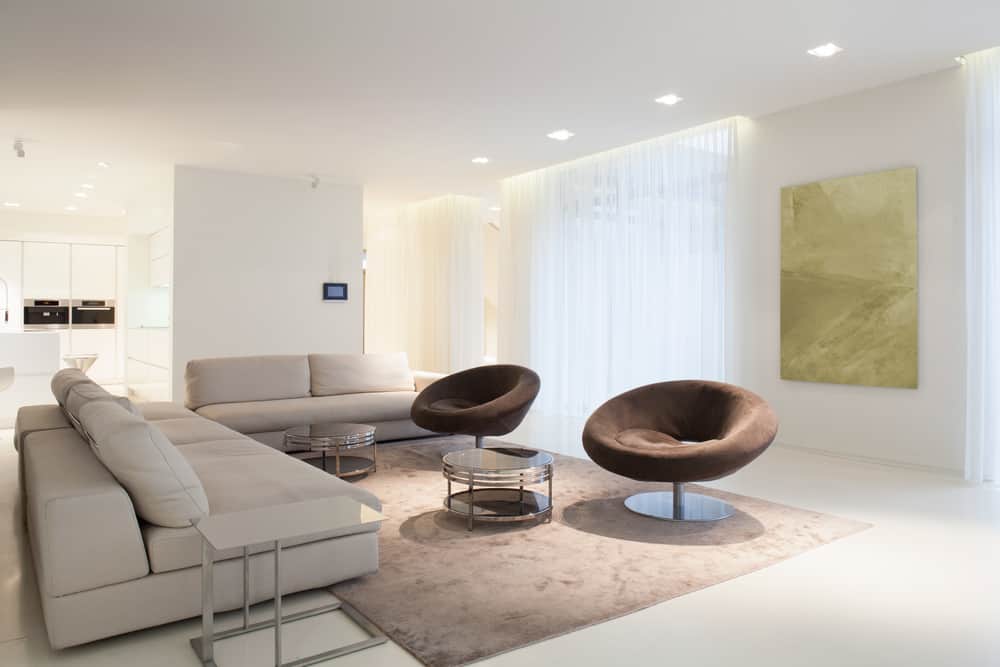 Modern minimalist living room with sleek beige sofas, brown swivel chairs, glass coffee tables, and soft ambient lighting