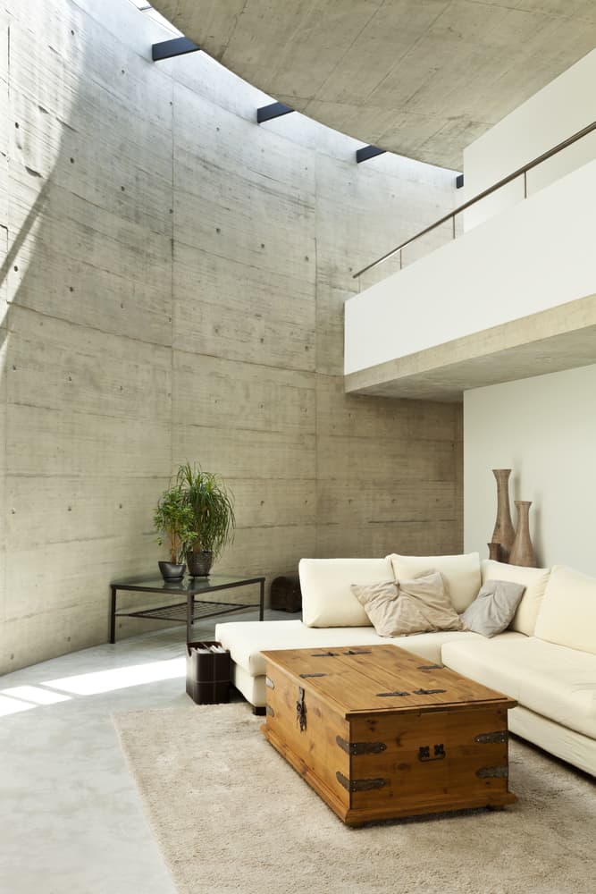 Brutalist-style living room with a curved concrete wall, cream sectional sofa, vintage wooden trunk, and natural lighting