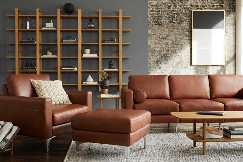 Modern living room with brown leather sofa, armchair, ottoman, and wooden bookshelf.