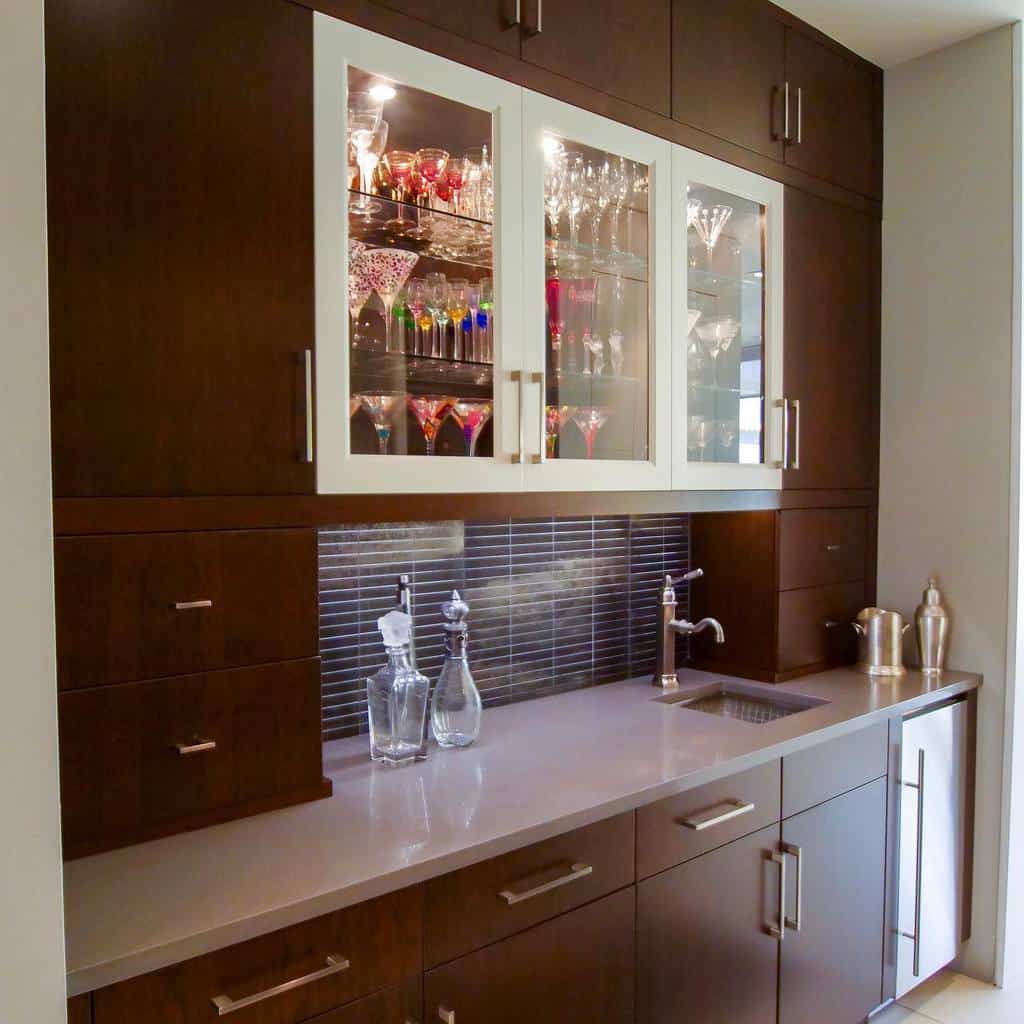 Sleek wet bar with dark wood cabinets, glass display, tiled backsplash, and compact sink.